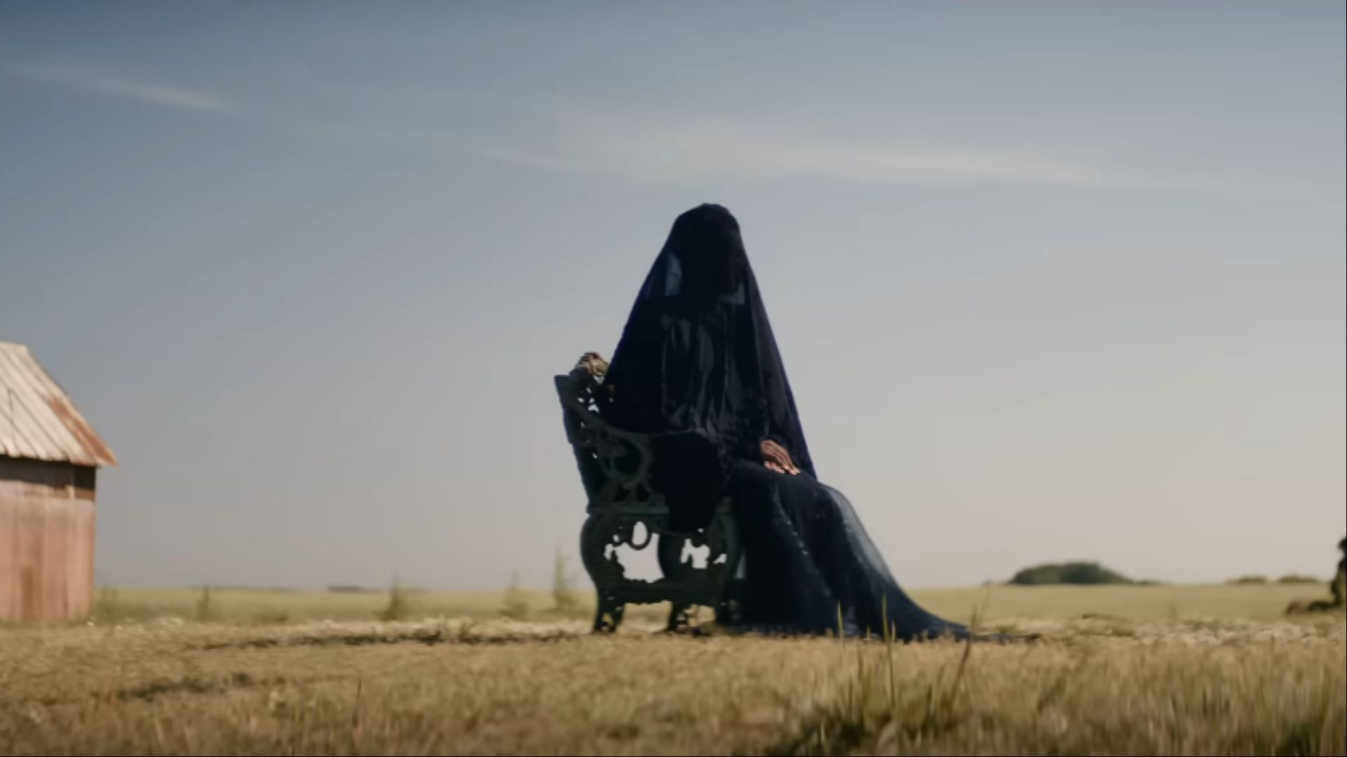 Seated in a wrought-iron chair, The Woman in the Yard waits under a blazing sun, her stillness more threatening than any movement (Image via Universal Pictures)